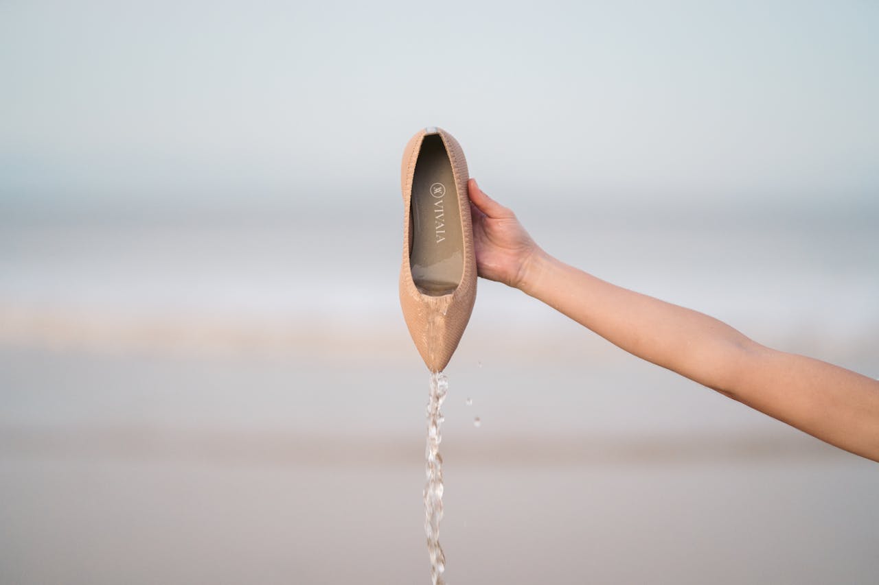 A fashionable beige shoe by the beach with water pouring out, held by a hand.